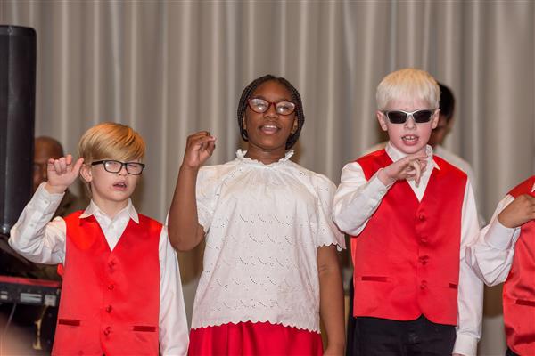 Three students smile widely while on stage singing a song. They are dressed in matching red and white. 